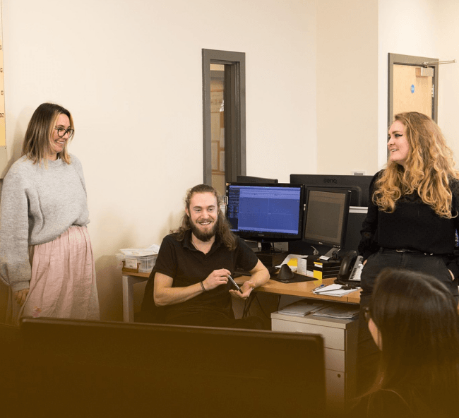 Three smiling team members in an office setting, collaborating near a computer workstation.