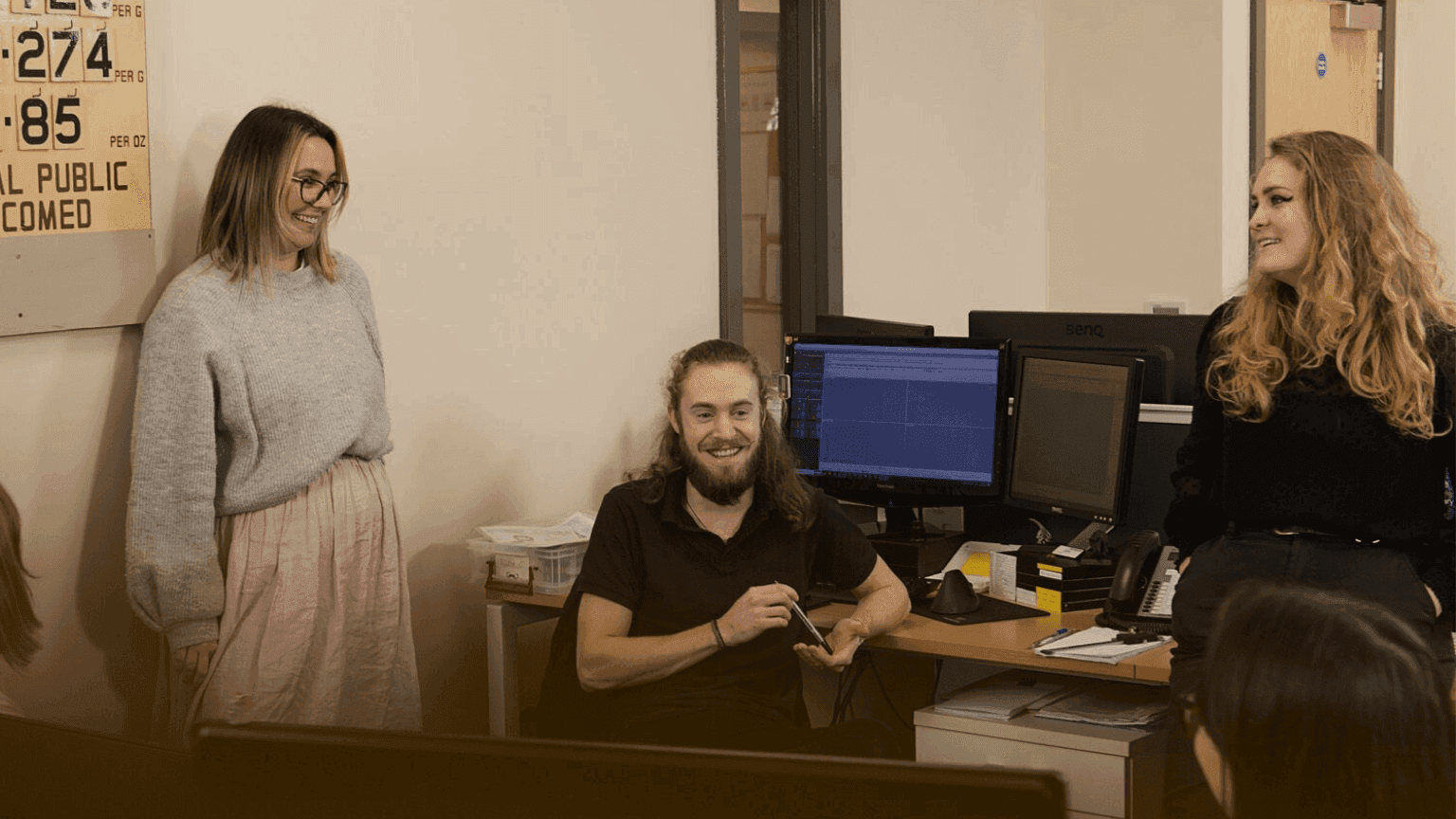 A small group of professional staff, two women and one man, collaborating and smiling at an office workstation.