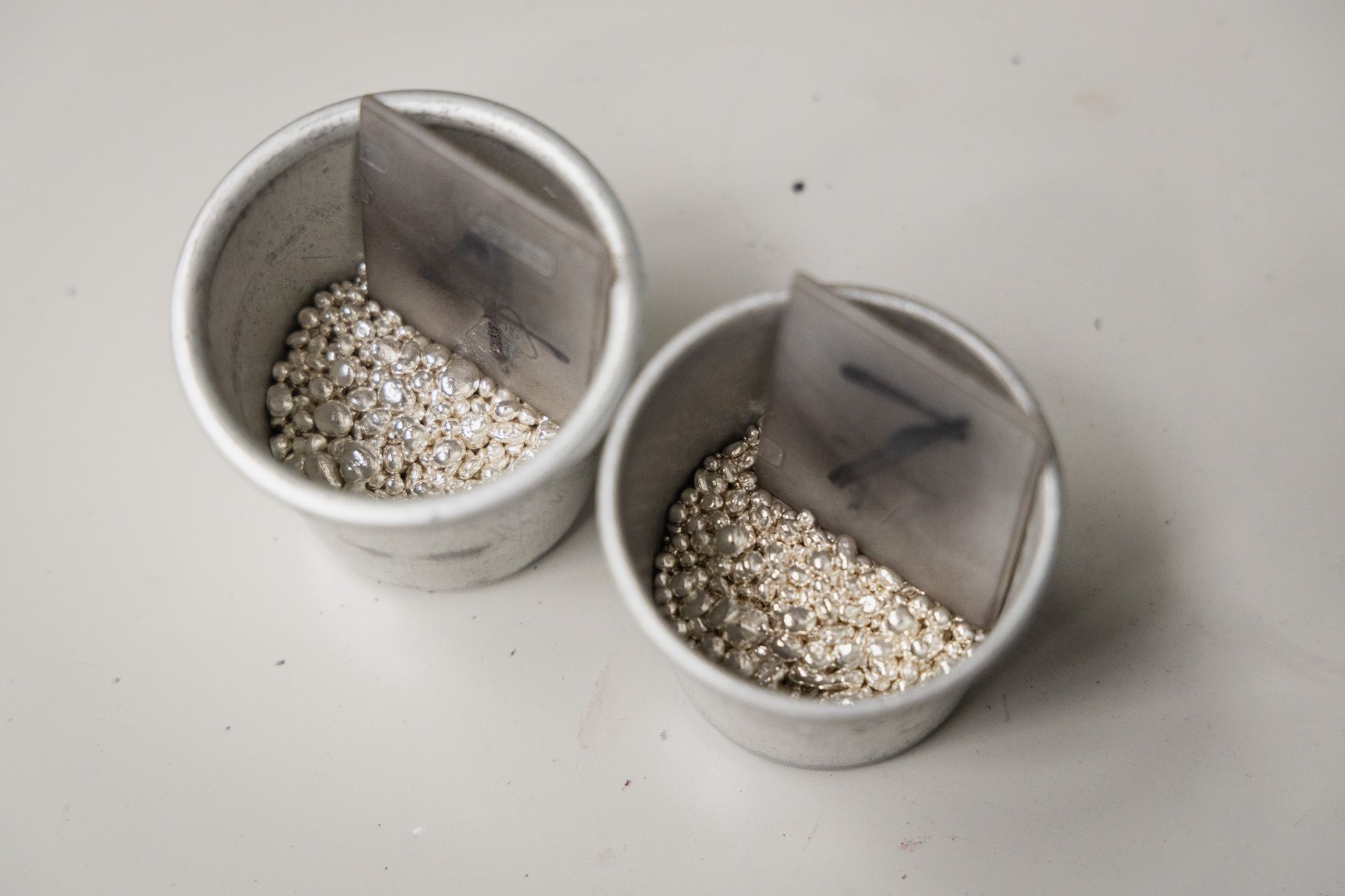 Two white metal cups, labeled "7" and "1", are shown from above, containing shiny silver-colored metal beads or shot, likely for jewelry casting or other industrial processes.
