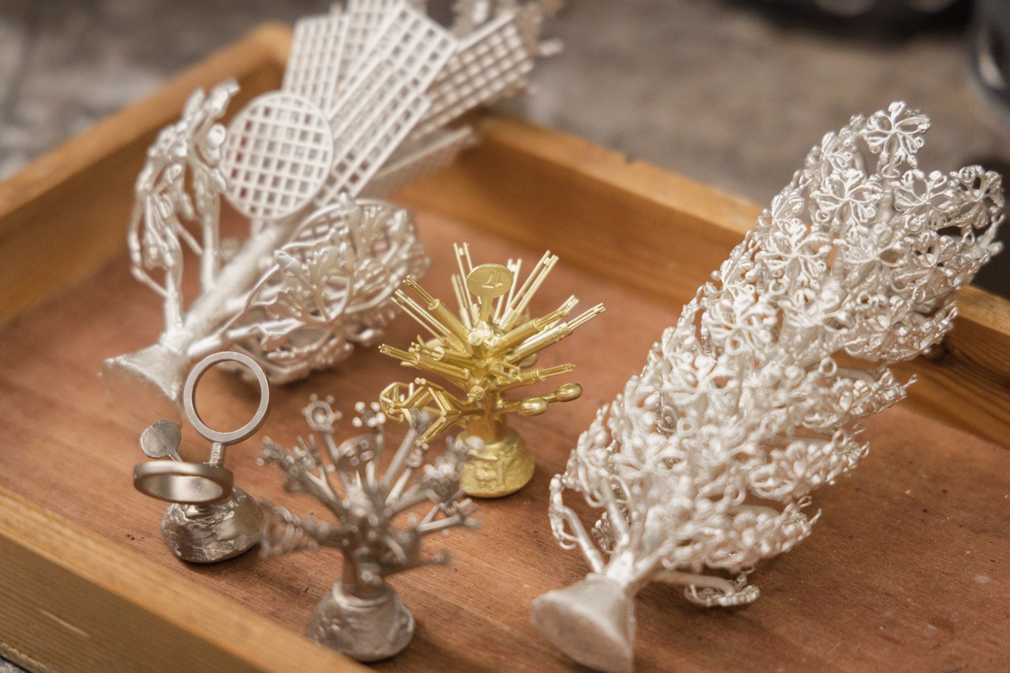 A close-up, elevated shot shows a wooden tray holding several metal casting trees, each with multiple rings and pendants attached.