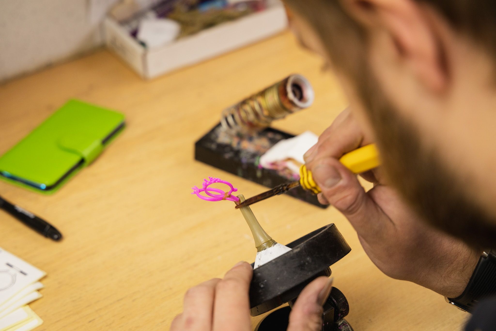 A jeweller with a beard works with a hot tool to carefully attach a pink wax ring model to a base.