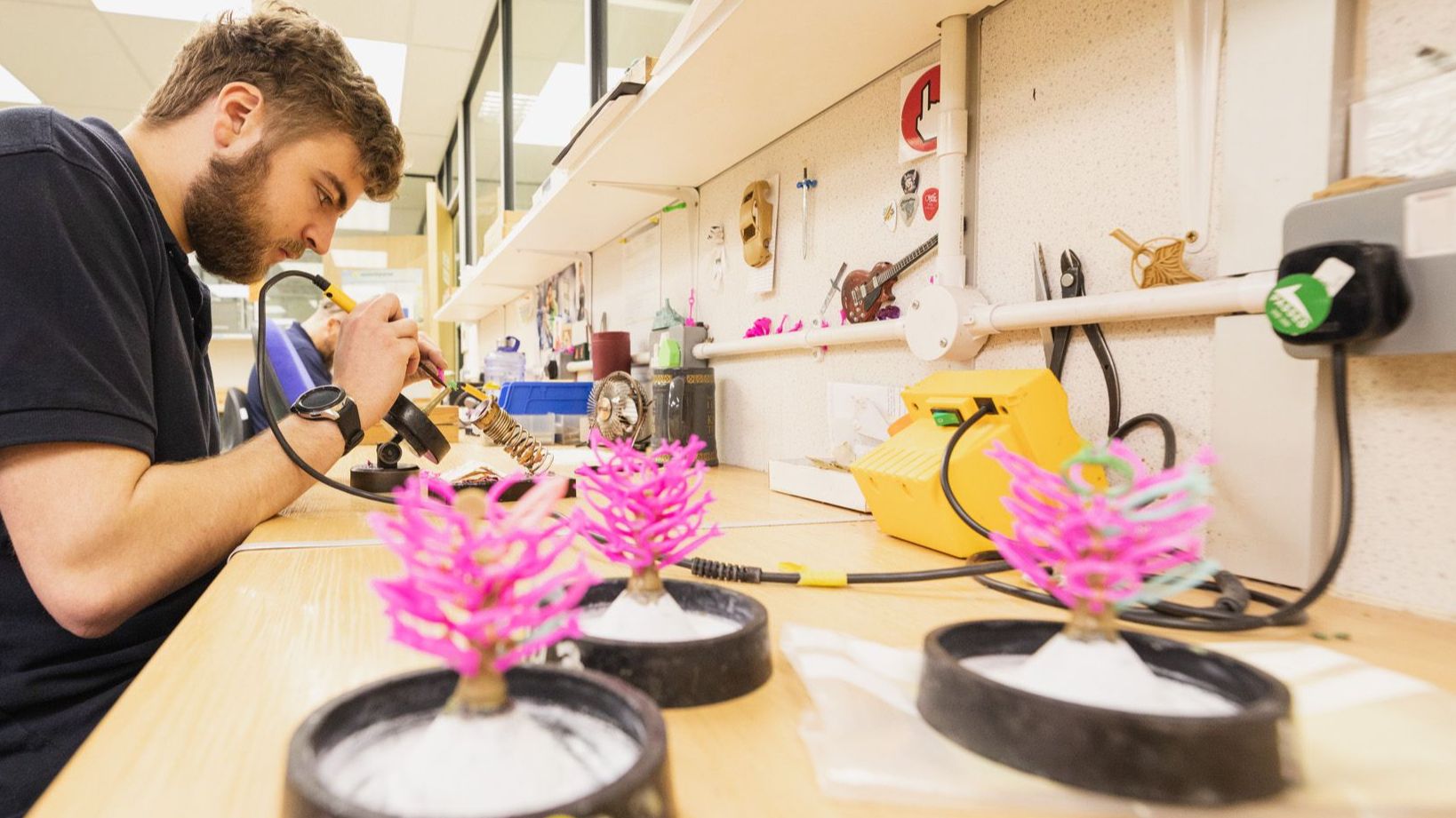 A jeweller with a beard is focused on soldering a piece of jewellery on a workbench, with pink wax 'sprue trees' in the foreground.