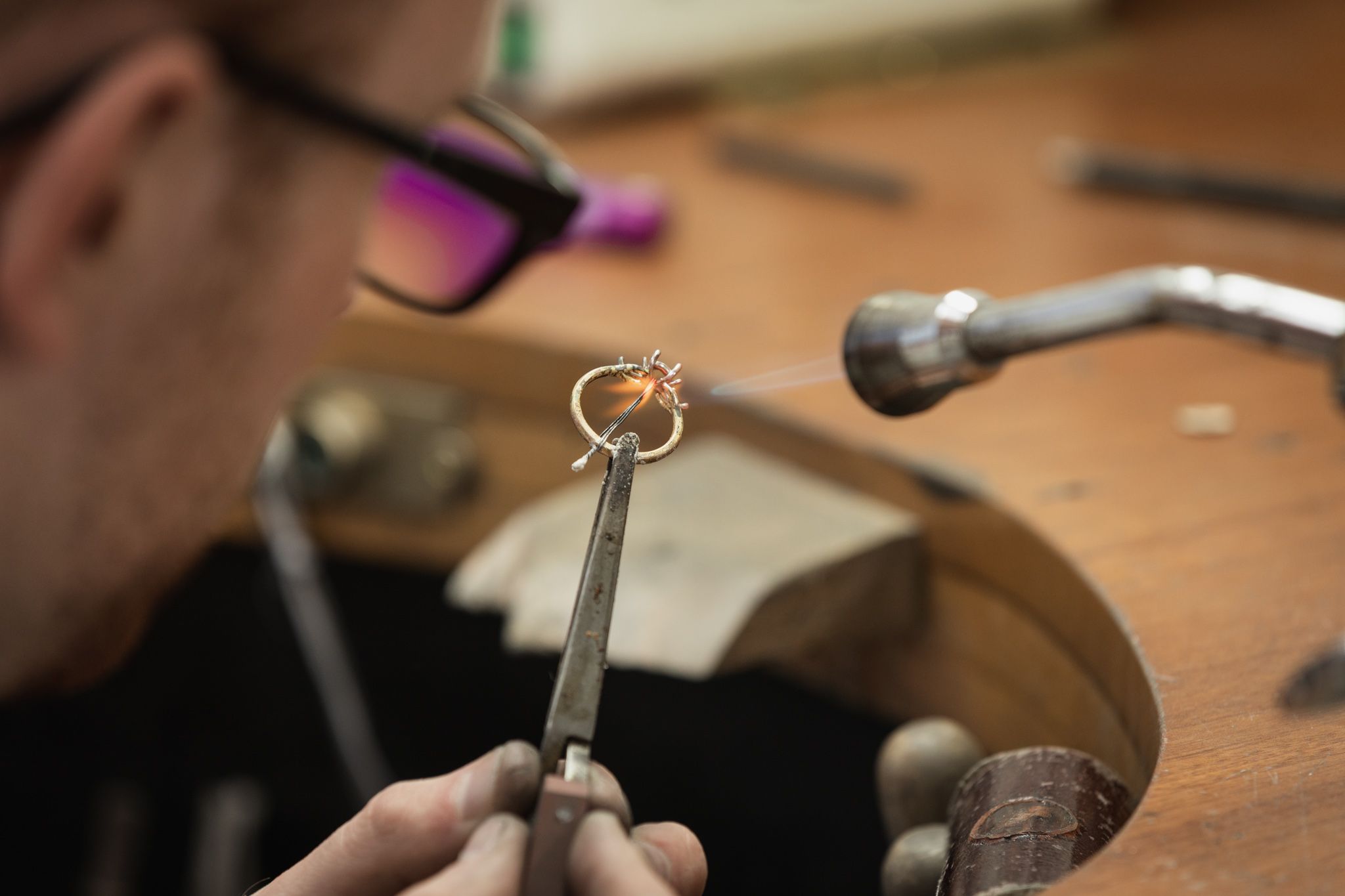 A close-up shows a jeweler wearing glasses, holding a gold ring with tweezers while using a small torch to solder a component, creating a bright orange flame.