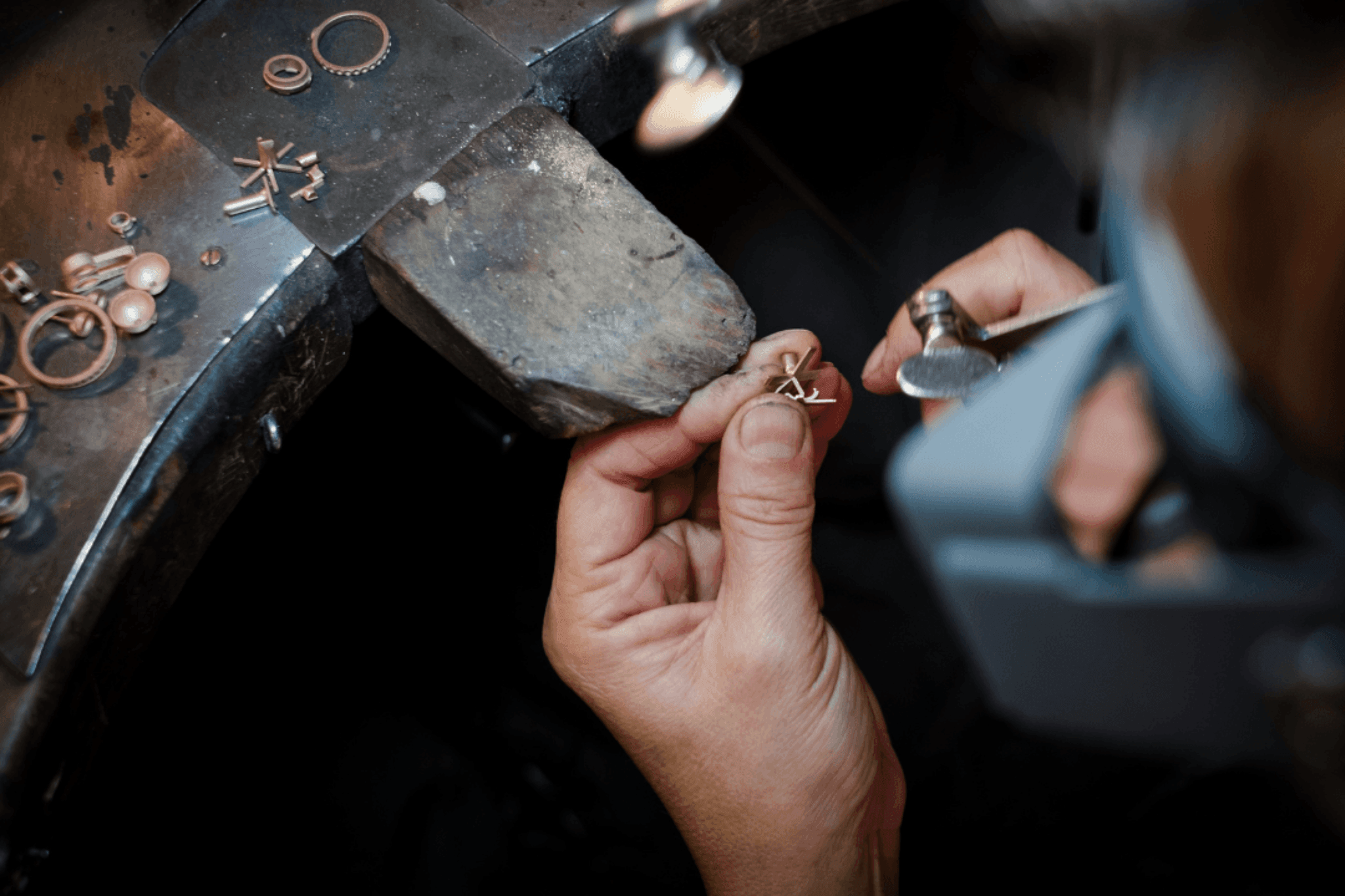 A close-up shows a person's hands meticulously working on a small piece of jewelry with tools, resting on a wooden jeweler's bench peg.