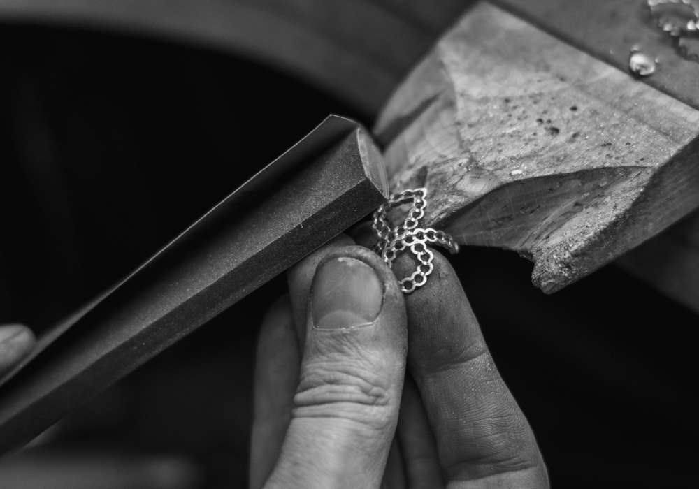 A close-up, black and white shot of a jeweller's hands holding a partially crafted, bespoke piece of jewellery. A tool is being used to shape the delicate metalwork.