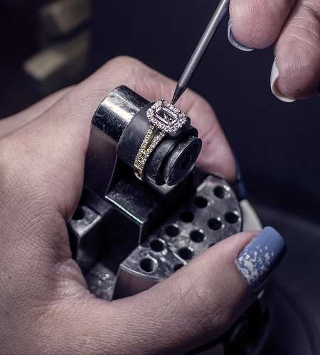  close-up shows hands carefully working on a ring set with a rectangular gemstone and surrounding smaller diamonds, held in a jeweler's vise.