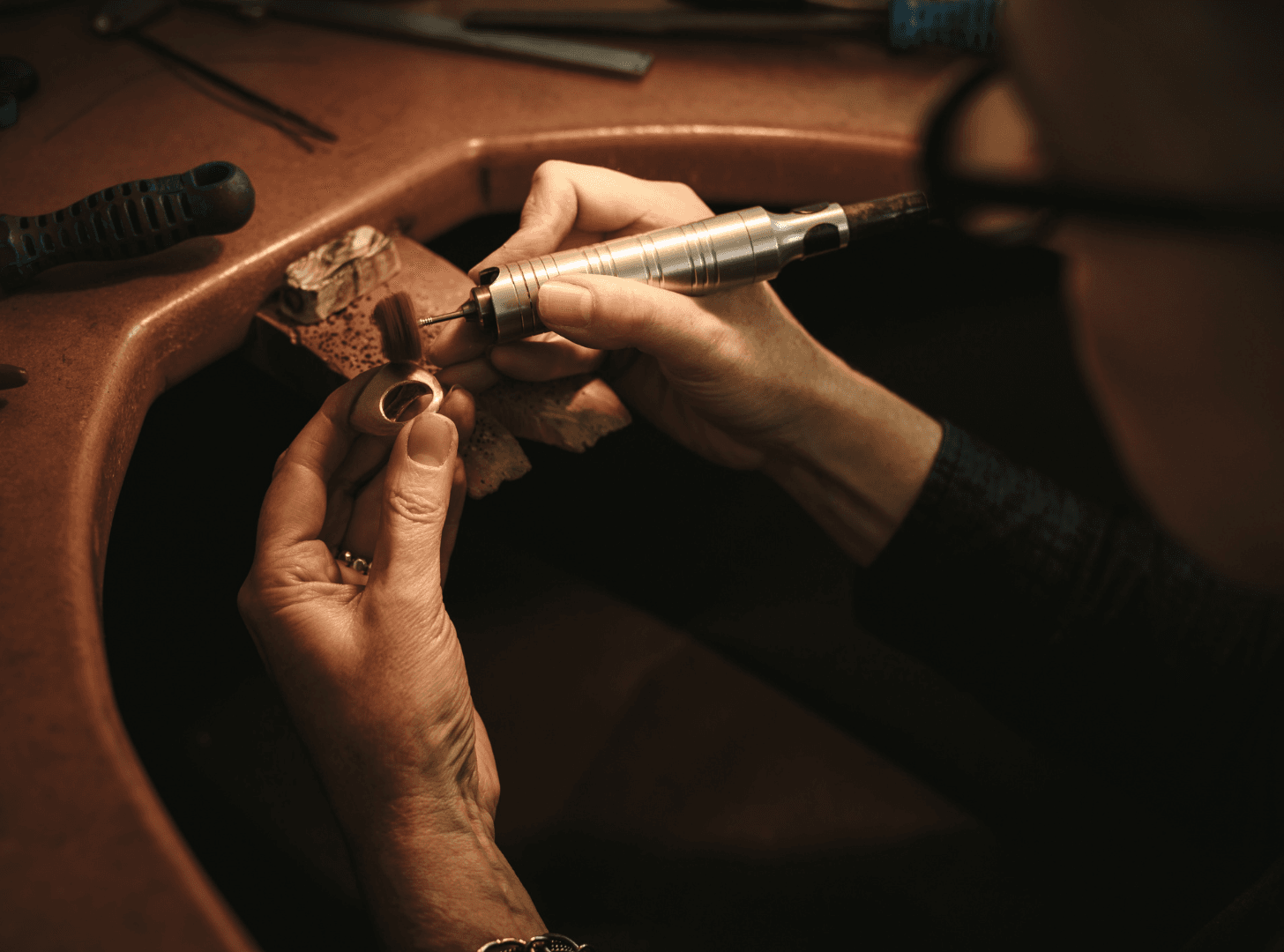A jeweler's hands meticulously work on a ring with engraving tools at a dimly lit workbench, conveying precision and craftsmanship.