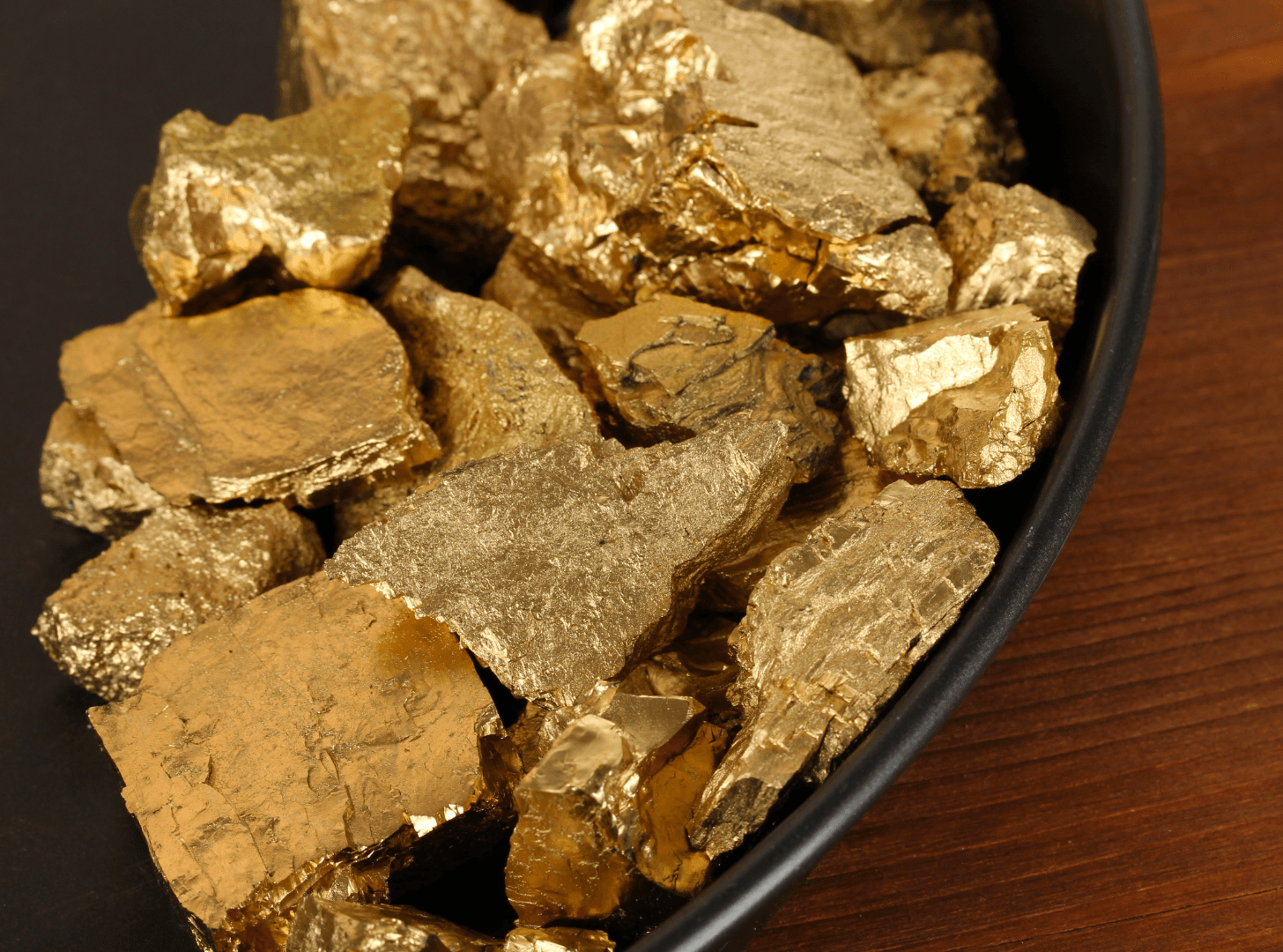 Close-up of shiny gold-colored mineral rocks, possibly pyrite or gold nuggets, displayed in a black bowl on a wooden surface.