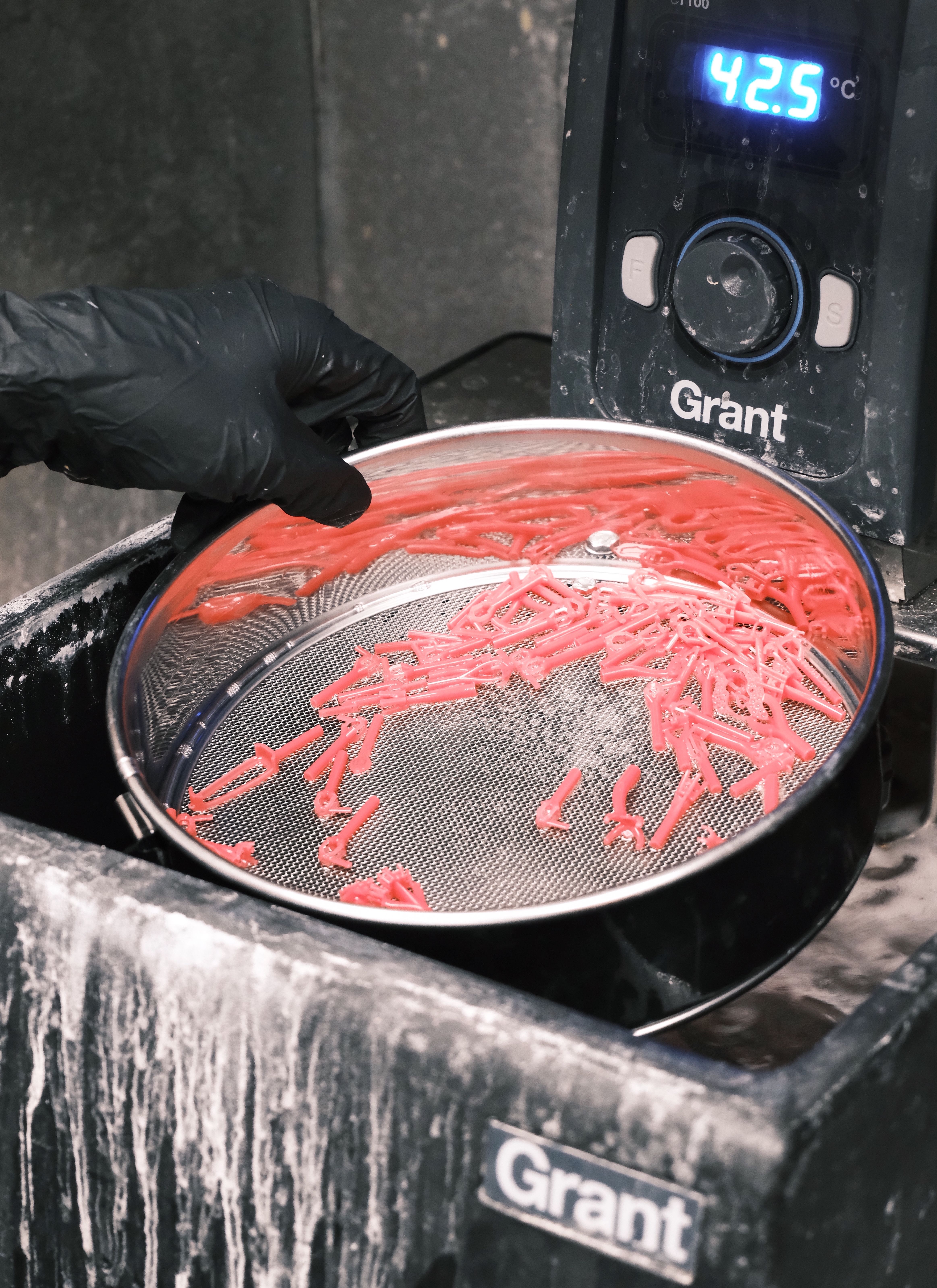 A gloved hand rinsing small red 3D-printed jewelry components in a sieve over a cleaning station displaying a temperature reading of 42.5 Degree's Celcius